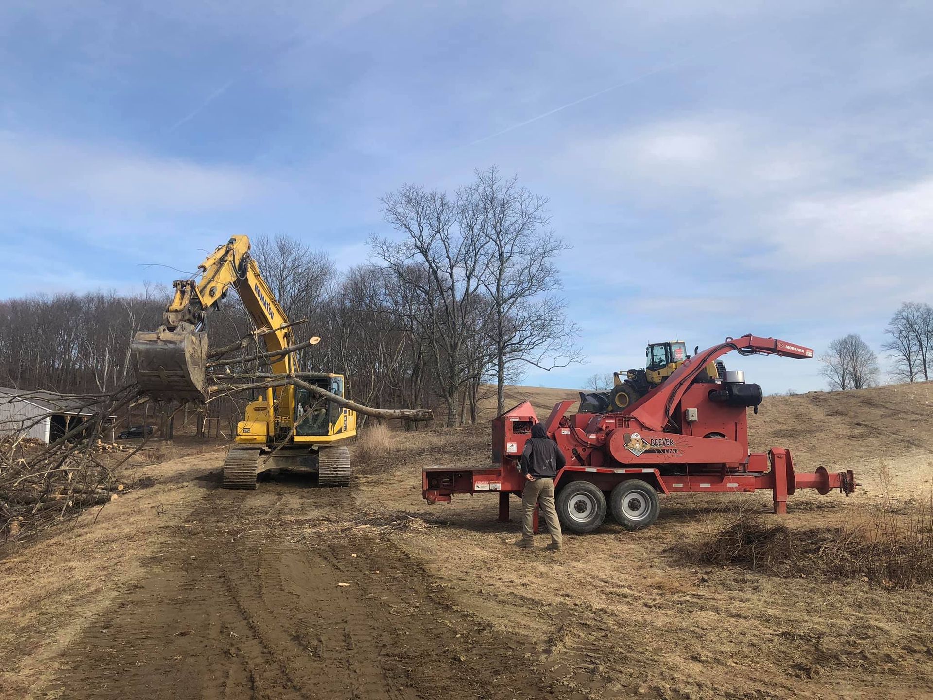 Excavator feeding branches to a wood chipper; a person stands nearby on a brown hillside with trees in the background.