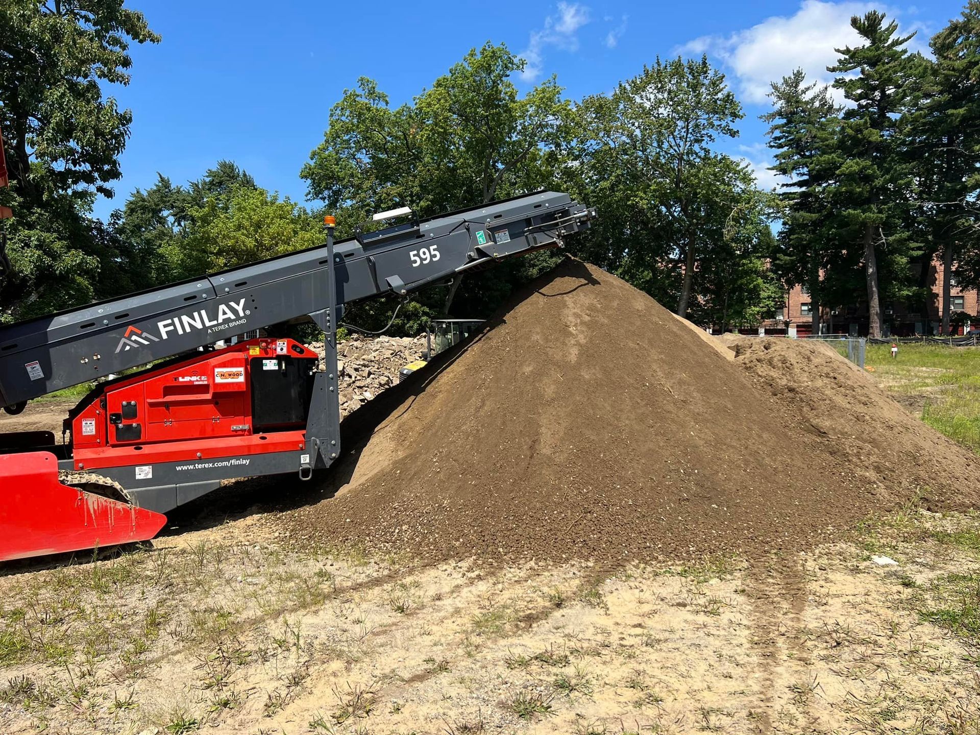 Red and black Finlay 510 mobile screener sorting dirt into a large pile in a grassy area under a blue sky.