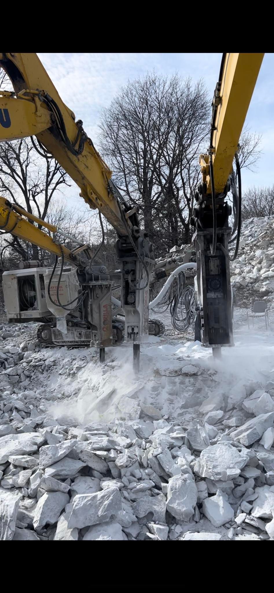 A large excavator demolishes a structure, breaking apart concrete, creating a cloud of dust.