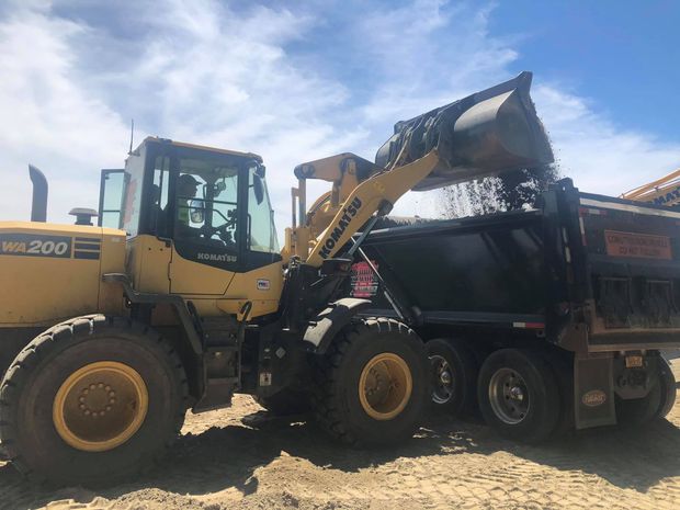 Yellow loader filling a black dump truck with gravel on a construction site under a blue sky.