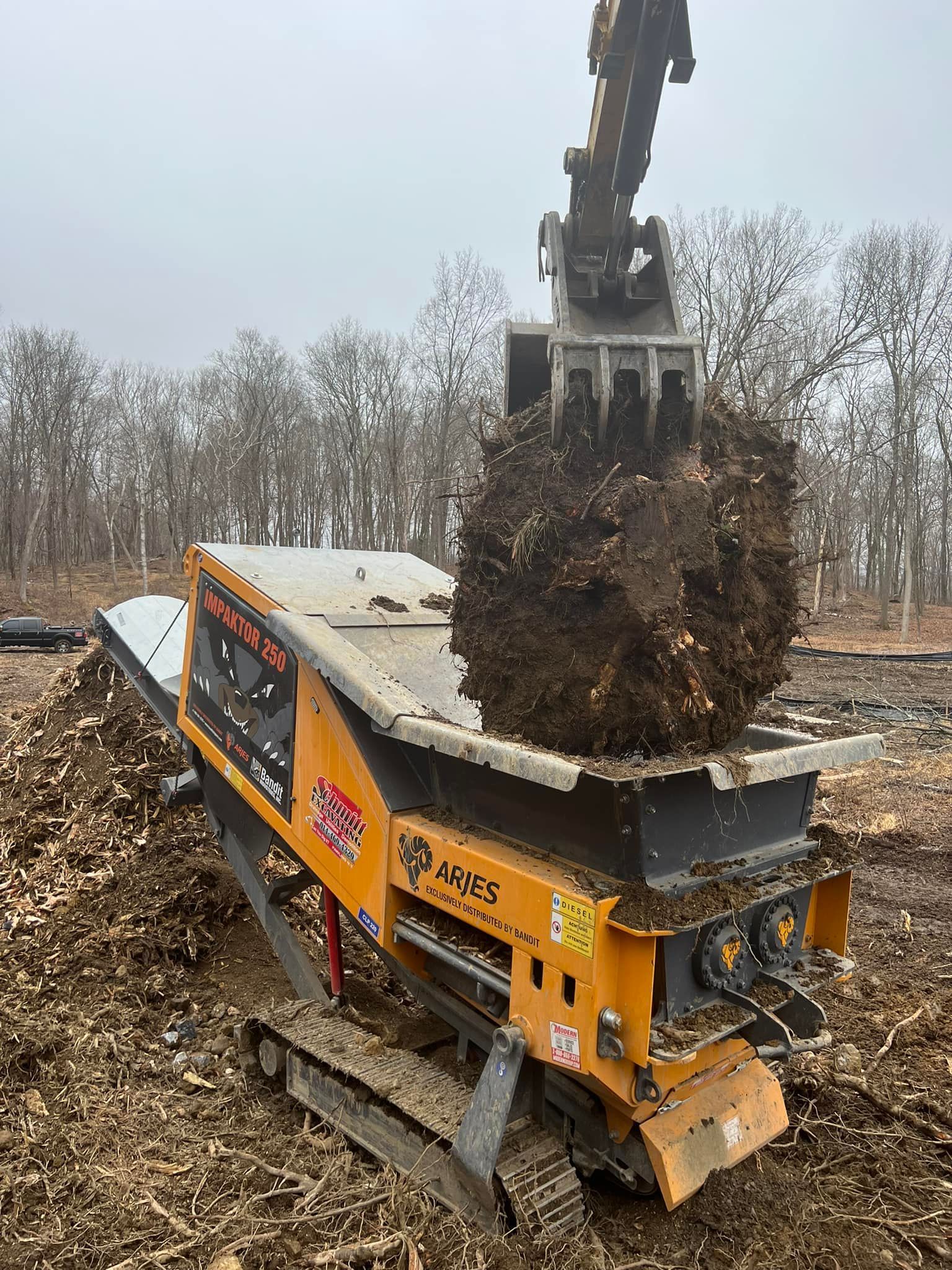 Yellow wood chipper receiving a load of wood from an excavator in a wooded area.