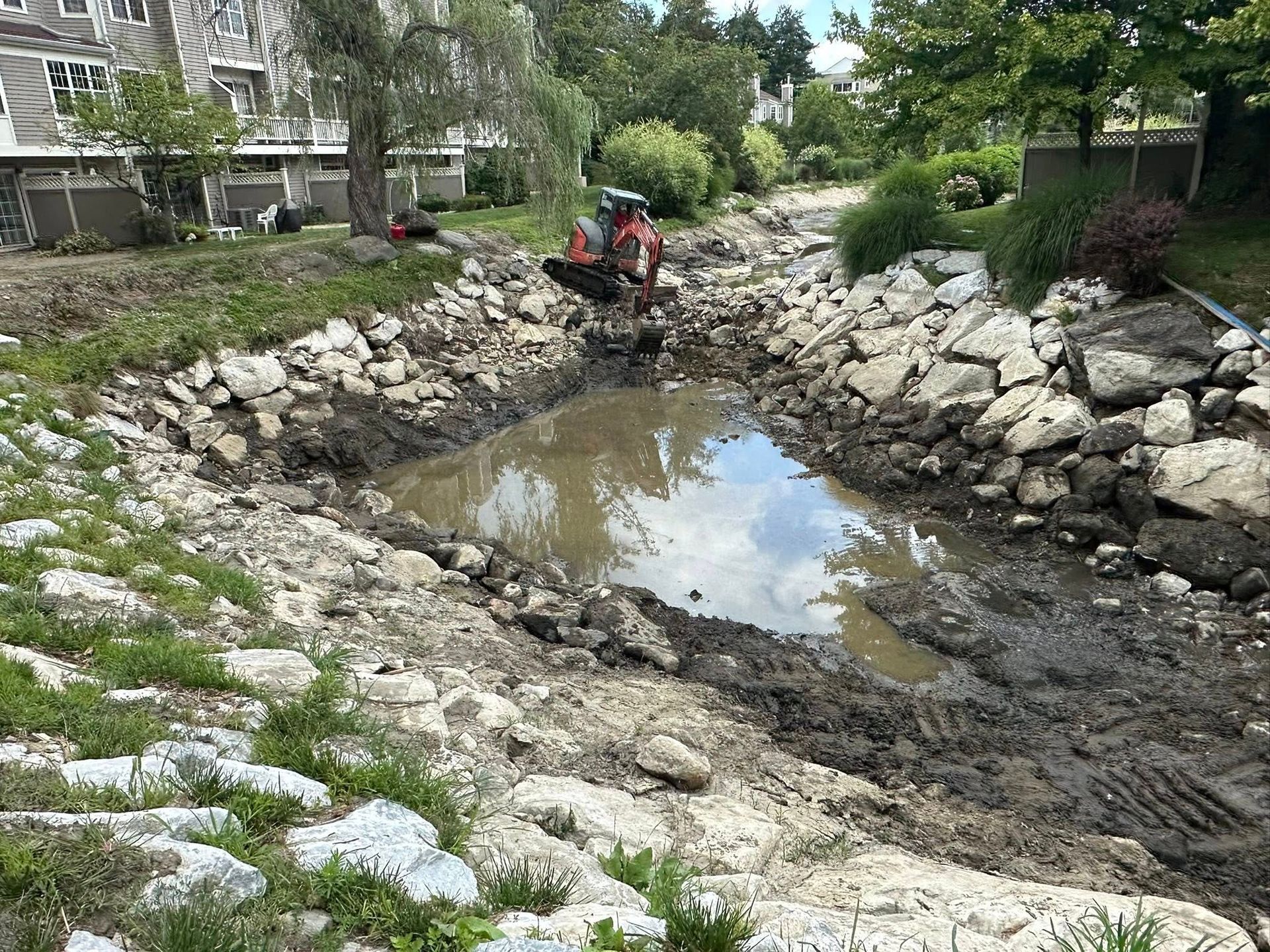 Construction of a small pond with rocks; an excavator works, mud and water visible, residential setting.