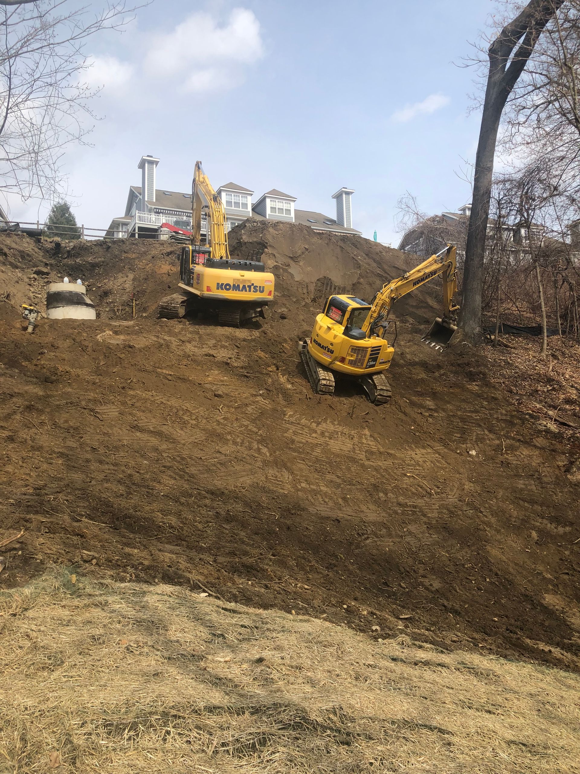 Two yellow excavators on a dirt hill. Buildings in the background under a blue sky.
