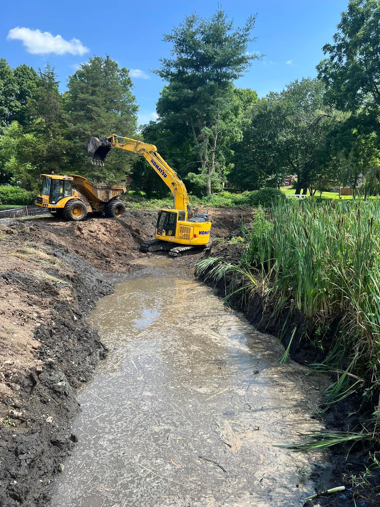 Yellow excavator and bulldozer clearing a muddy ditch near trees on a sunny day.