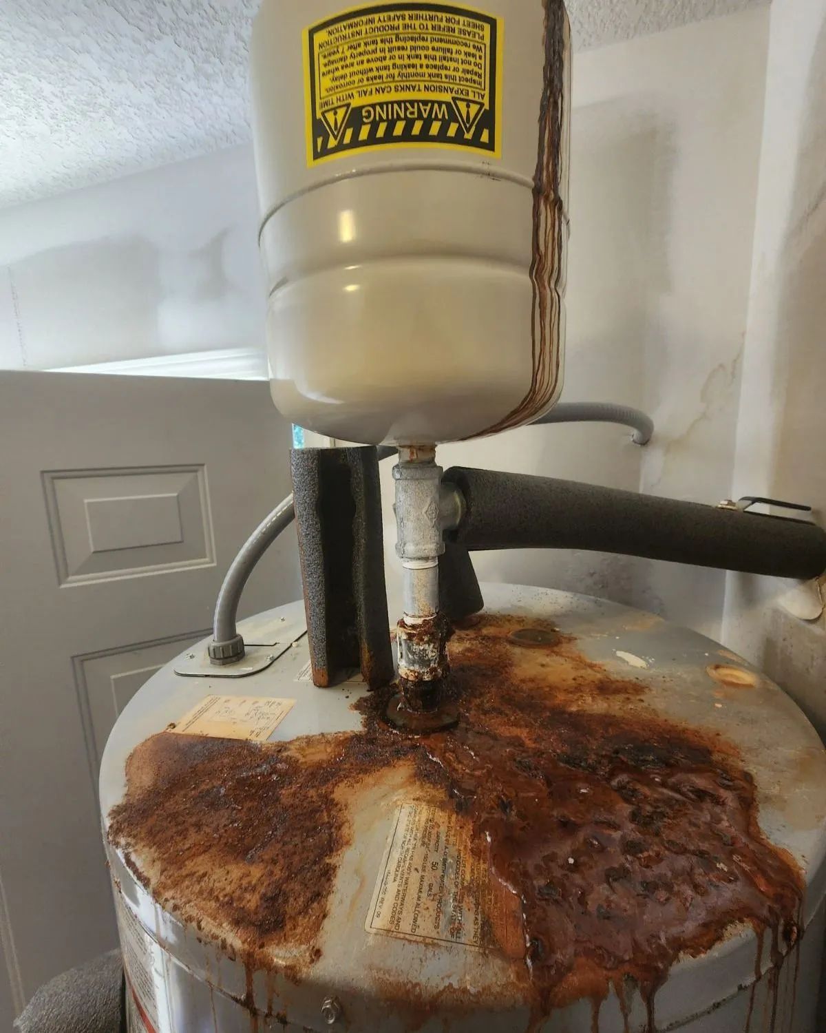 Rusty water heater with heavy corrosion and stains around the top and pipes in a utility room