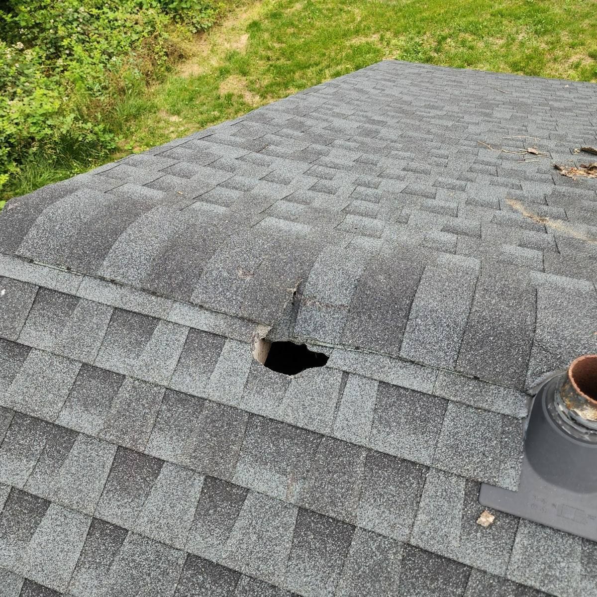 Damaged asphalt roof shingles with a hole near the ridge and a vent pipe on the right