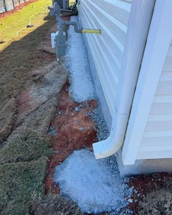 Side of a building with a downspout draining onto gravel and red soil beside a water spigot.