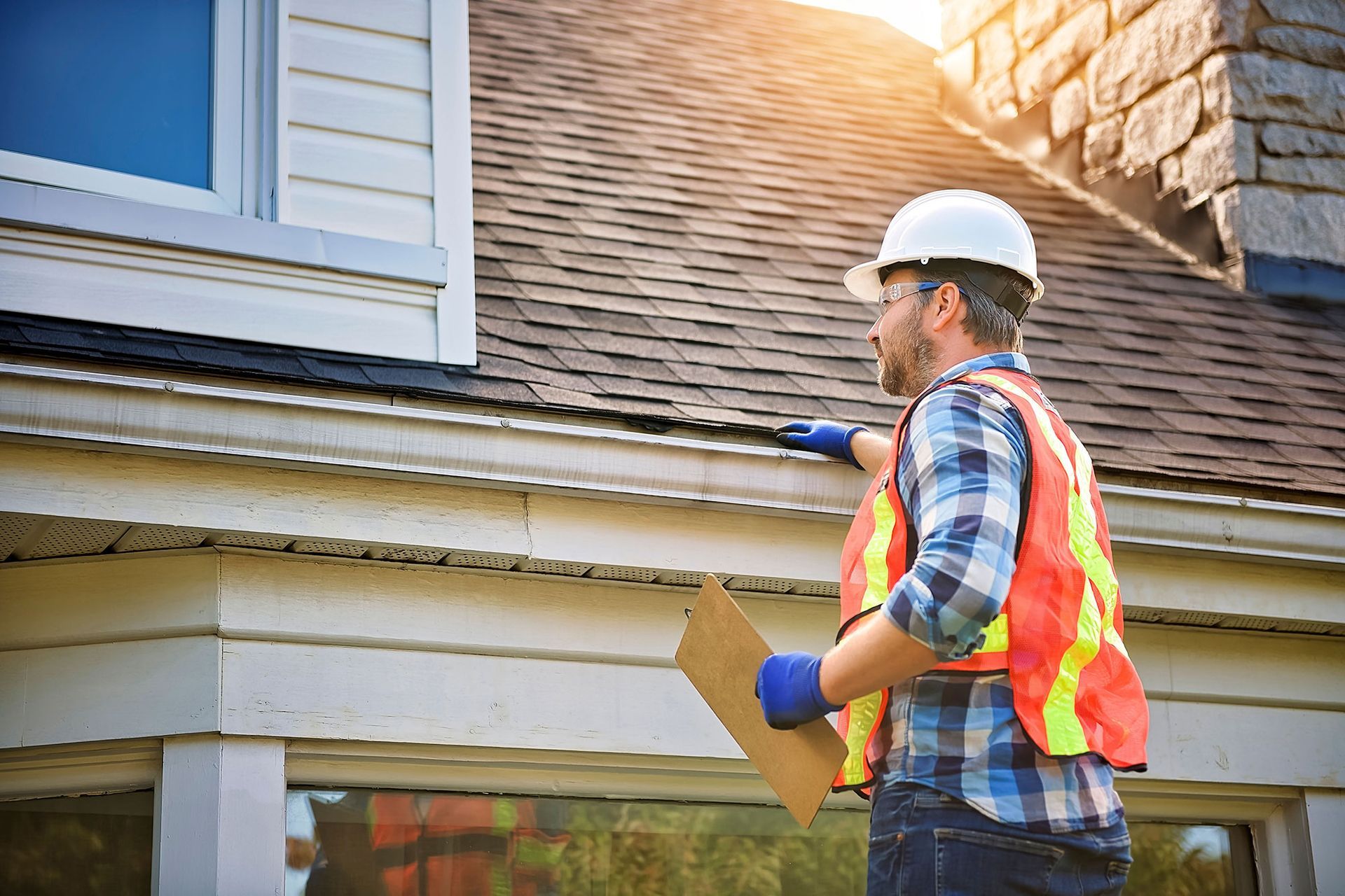 Inspector in hard hat and safety vest examining a house roof with a clipboard
