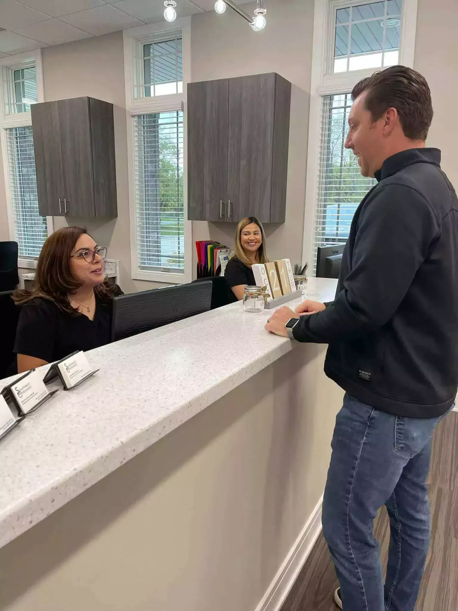 Two people at a reception counter in a modern, well-lit office; one person stands while the other sits and speaks.