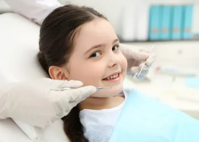 A smiling child sits in a dental chair while a dentist wearing gloves prepares an examination tool.