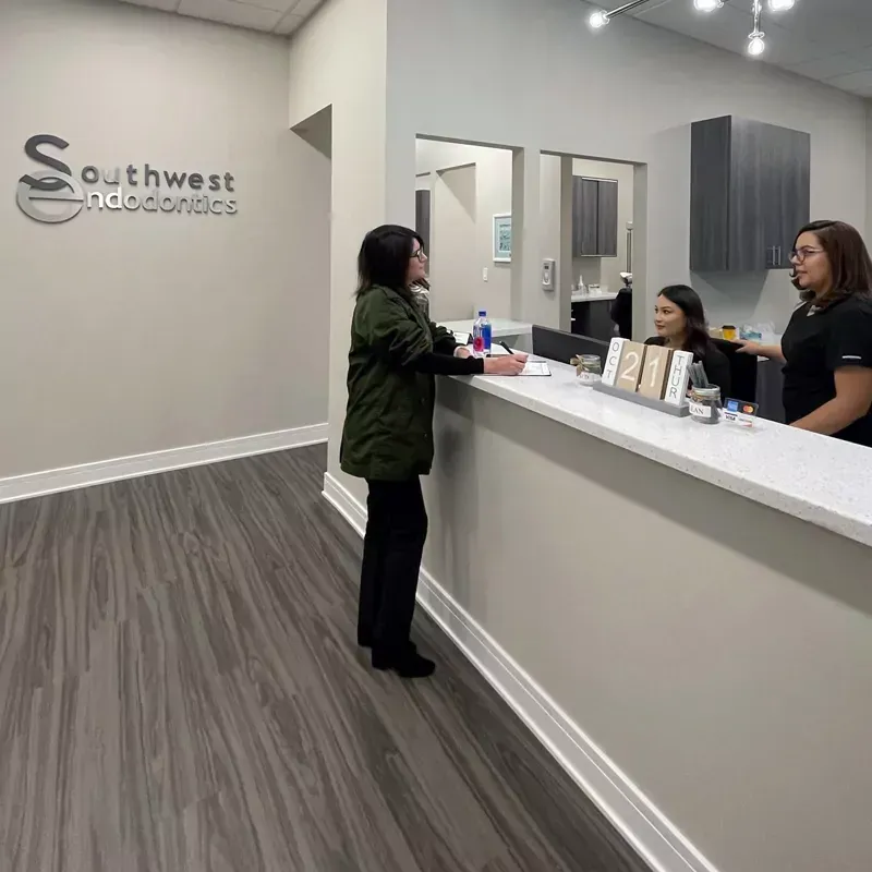 A person stands at a reception desk signing papers while two staff members assist them in a dental office.