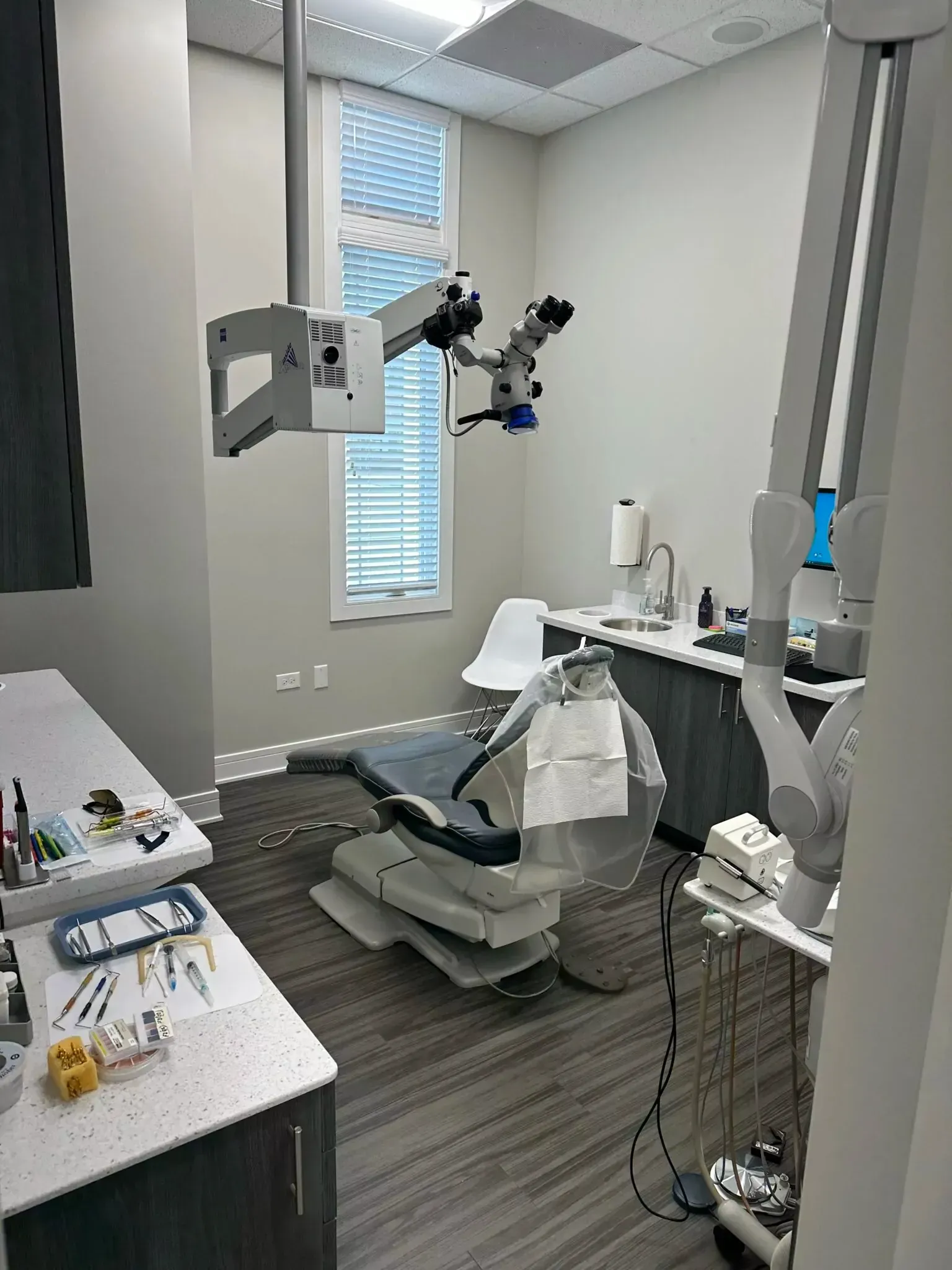 Modern dental office with a treatment chair, specialized overhead microscope, sink, and dental instruments on a counter.