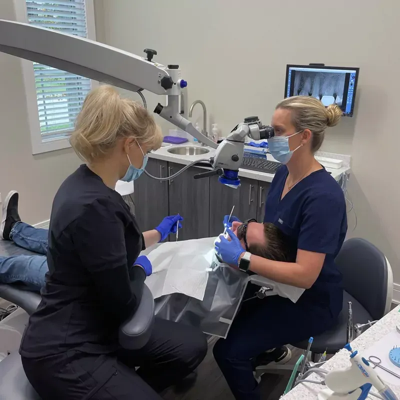 Two dental professionals wearing masks and blue gloves perform a procedure on a patient using a dental microscope.
