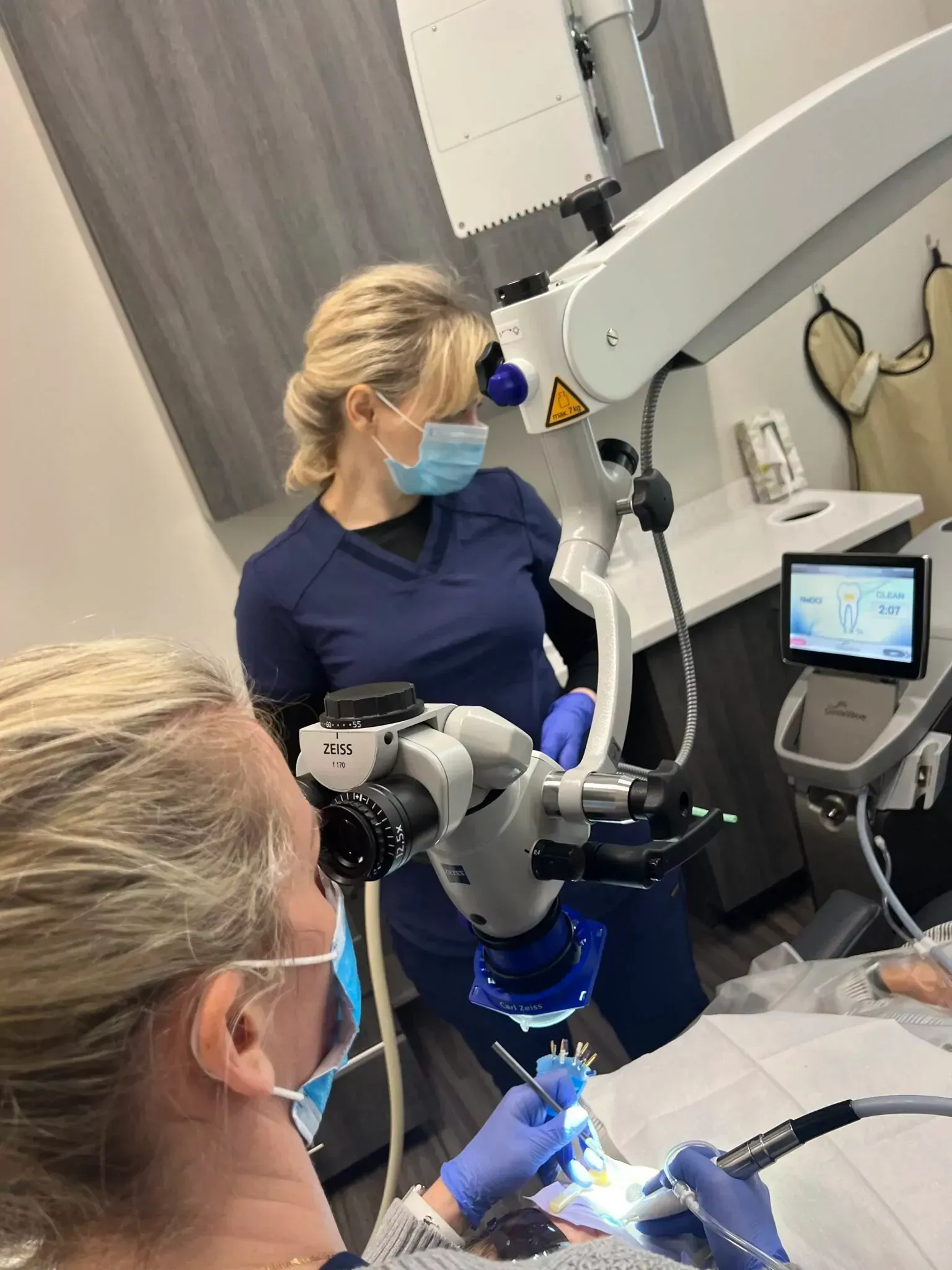 Two professionals in medical scrubs and masks perform a procedure using a high-magnification dental microscope.