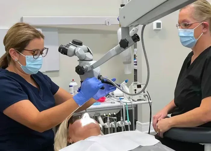 A dentist uses a microscope to perform a procedure on a patient in a clinical setting, with an assistant observing.