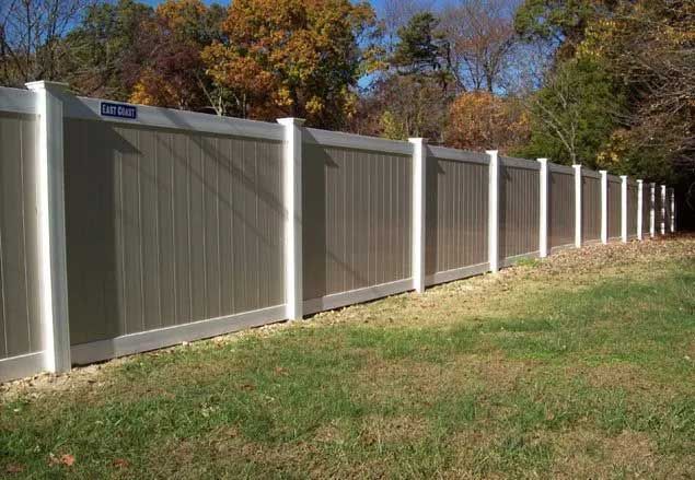 Beige vinyl fence with white posts in a grassy yard, trees in the background.