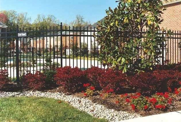 Black metal fence surrounding a landscaped yard with red bushes and flowers.