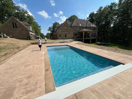 A rectangular swimming pool with turquoise water and stamped concrete surround, near a stone house and trees.
