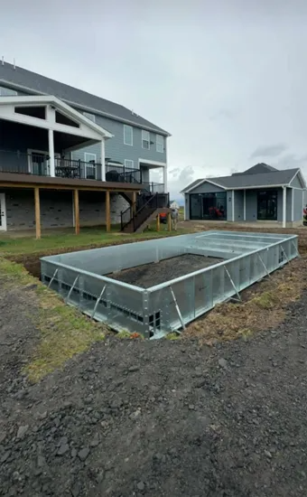 Partially built rectangular pool next to a two-story gray house and small outbuilding under an overcast sky.