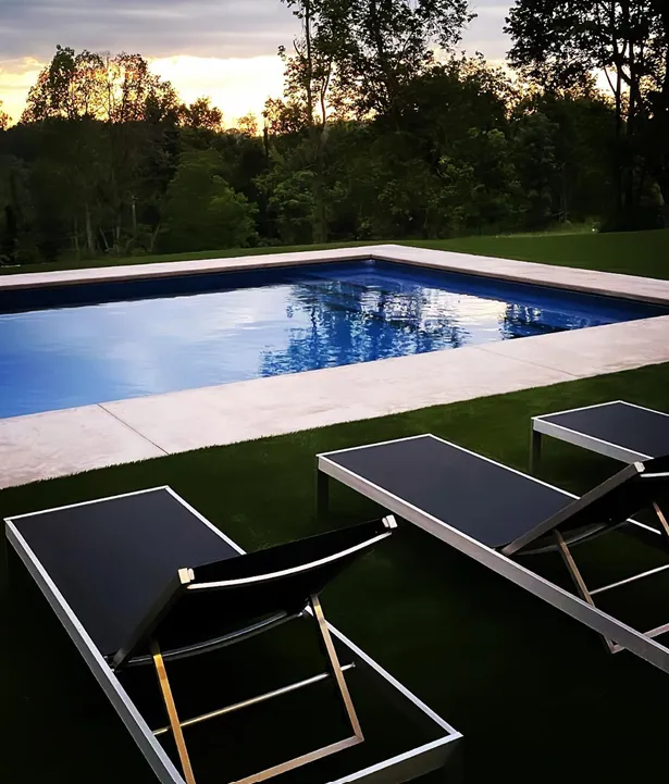 Swimming pool with black lounge chairs on green grass; trees in the background under a cloudy sky.