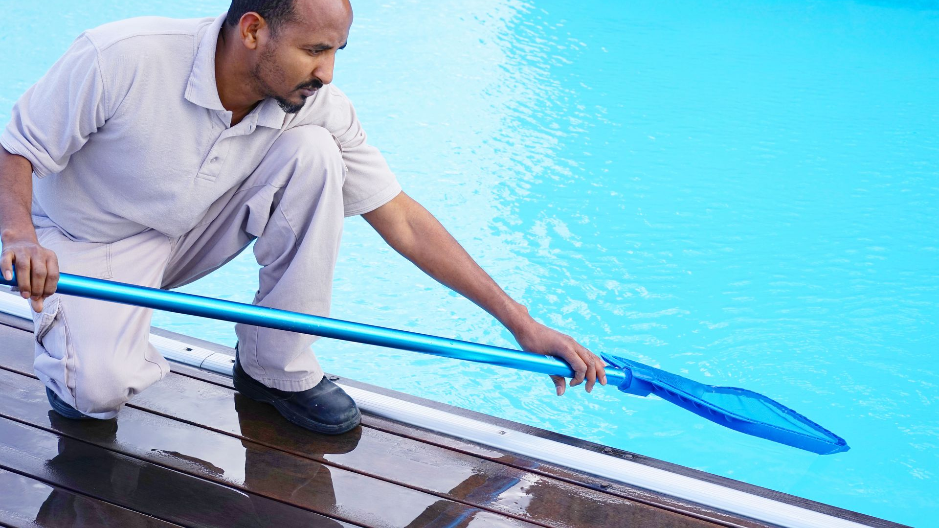 Man using a pool skimmer to clean a swimming pool. He is kneeling on a wooden deck.