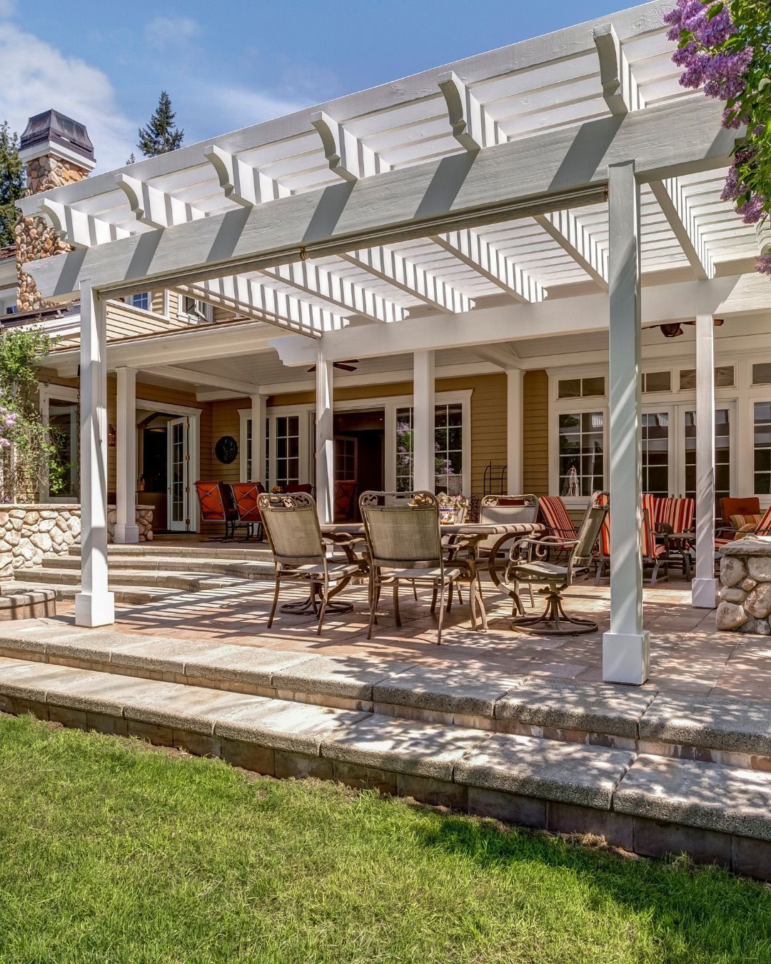 Patio with pergola, outdoor dining set, stone steps, and green lawn in sunlight.