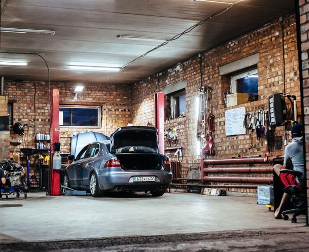 Car in a garage with open trunk, raised on a lift. Red brick walls, tools, and a person seated to the right.