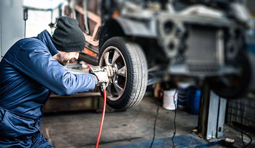 Mechanic in blue jumpsuit removes car tire with an impact wrench in a garage.
