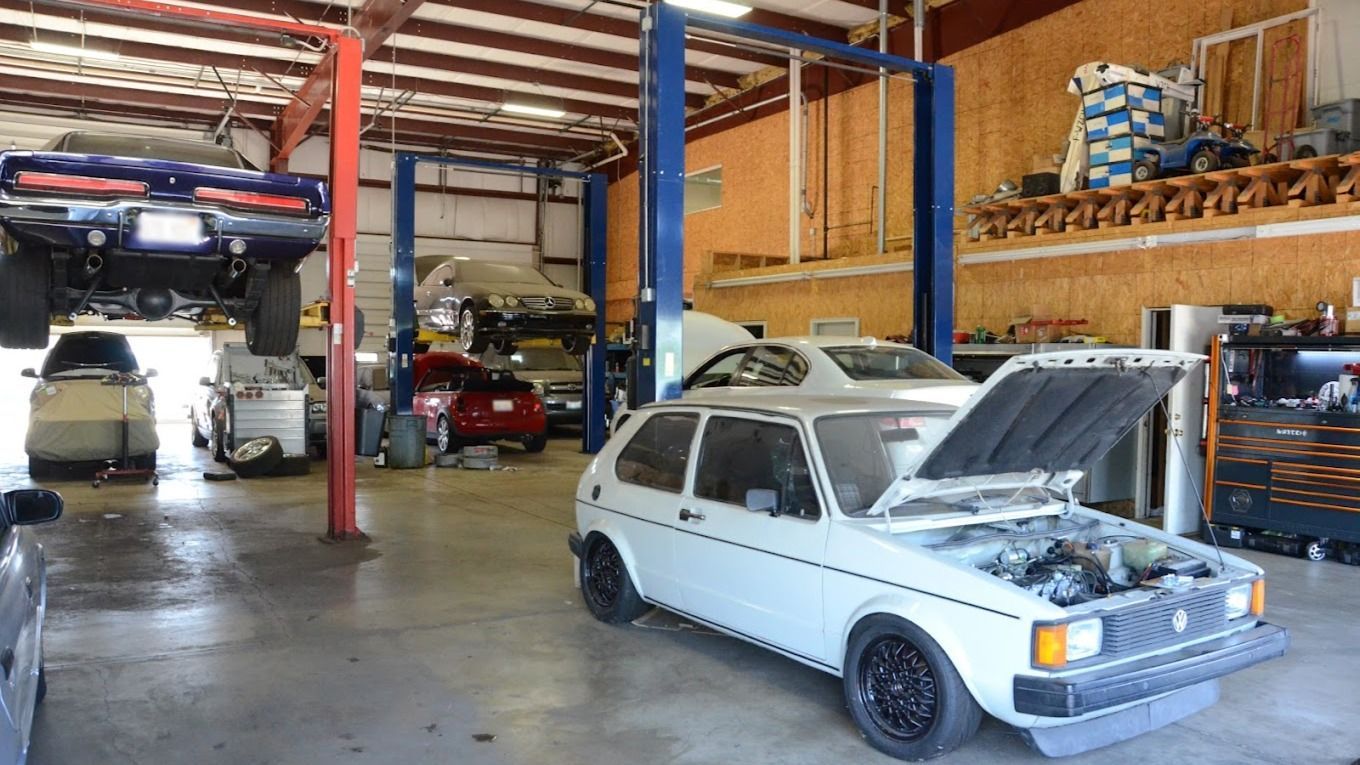 Auto repair shop, cars on lifts, mechanics working. White car with hood open in foreground.