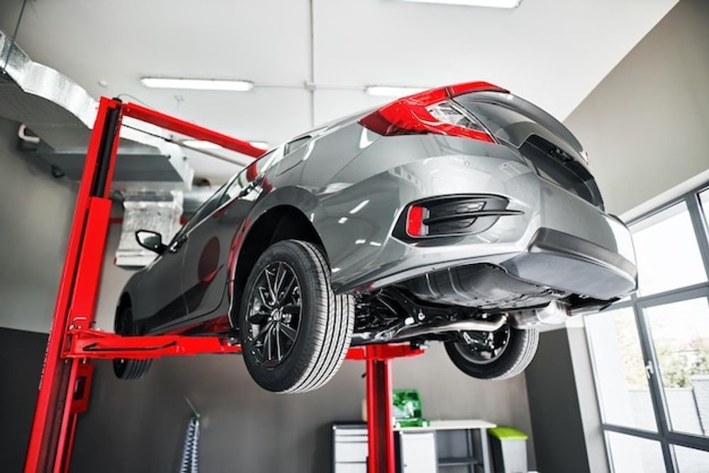 A grey car raised on a red car lift inside a brightly lit auto repair shop.