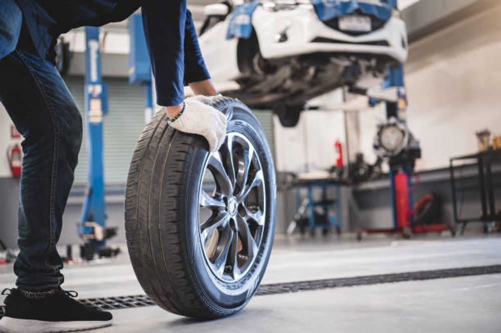 Mechanic holding a car tire in a repair shop with a car on a lift.