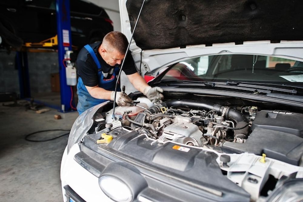 Mechanic working on a car engine in a garage. Car hood is open; the mechanic is wearing overalls.