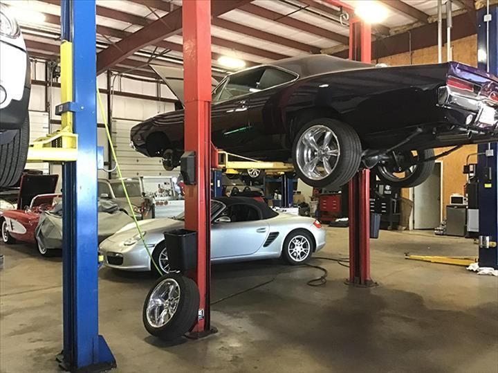 Cars on lifts in a garage; a classic dark car is elevated on red posts; a silver car and tire are on the floor.
