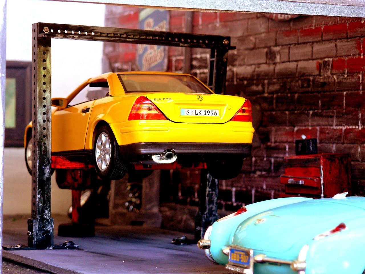 Yellow car raised on a lift in a garage, next to a blue car. Brick wall backdrop.