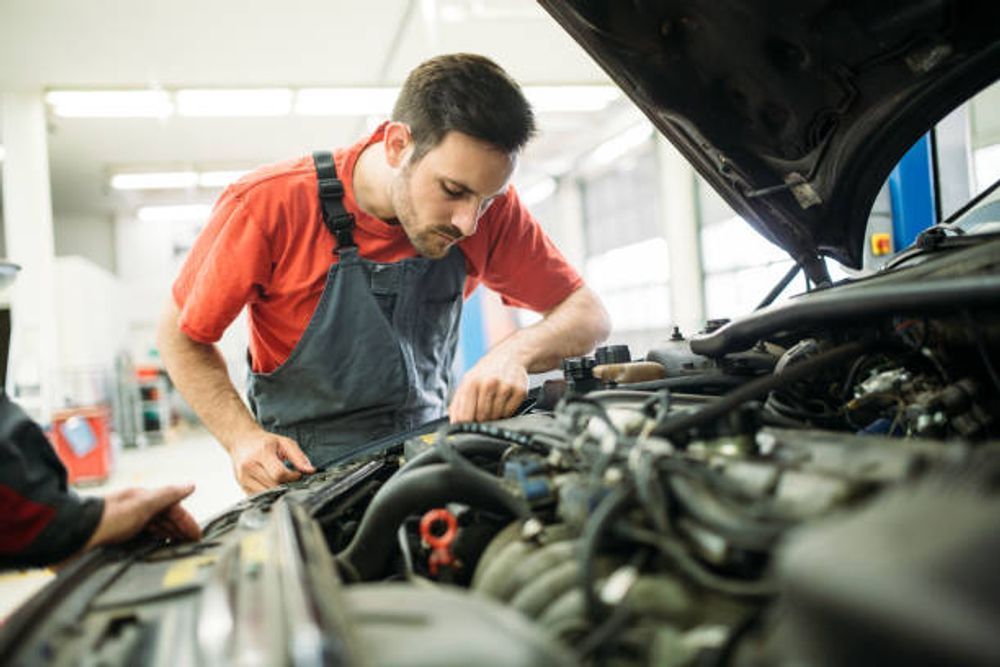 Mechanic in red shirt and blue overalls works on a car engine in a garage.