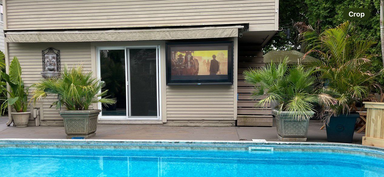 A swimming pool in front of a house with a television mounted on the wall.