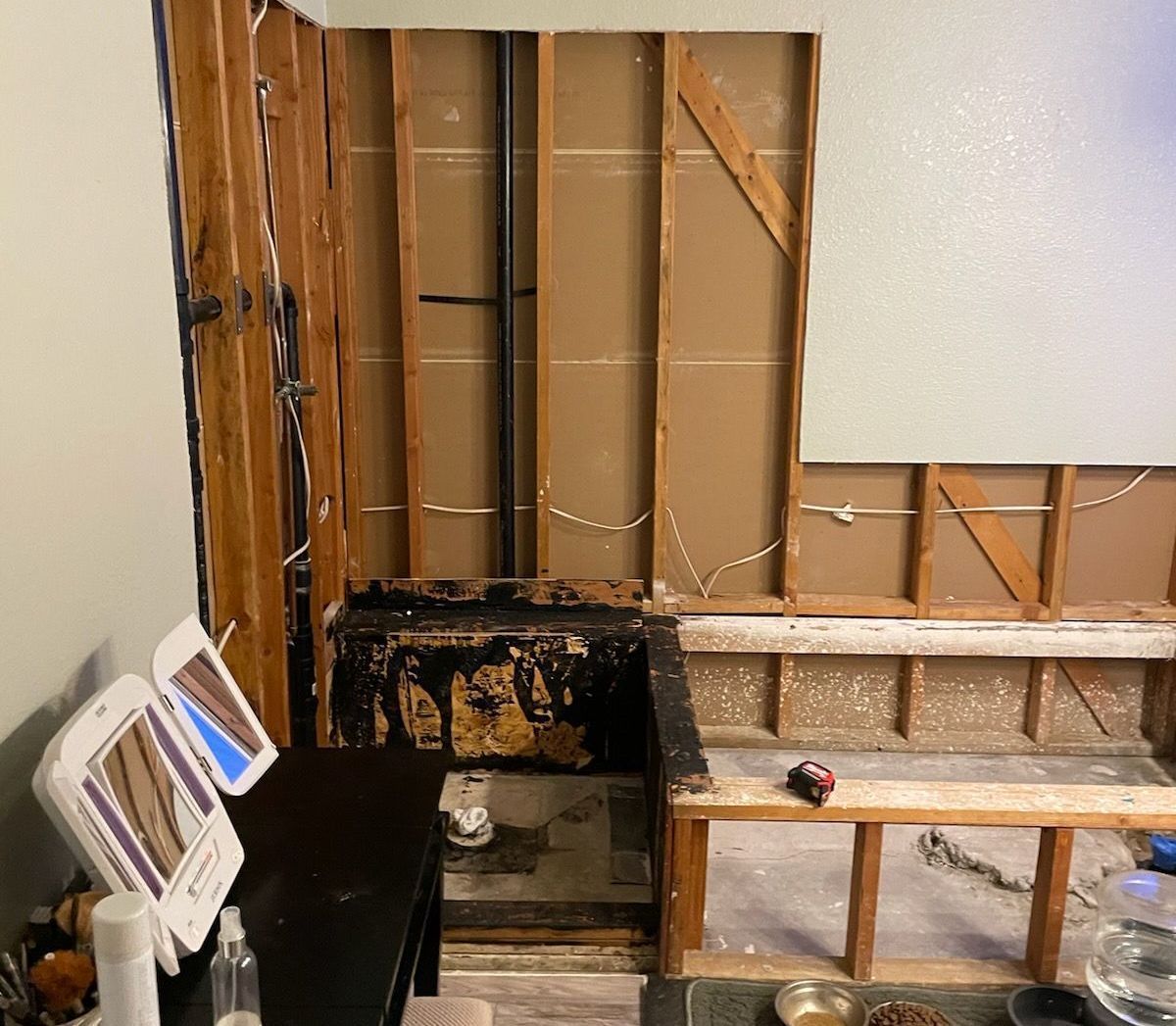 Bathroom wall under construction, with exposed framing, pipes, and damaged flooring; a vanity and mirror are visible.