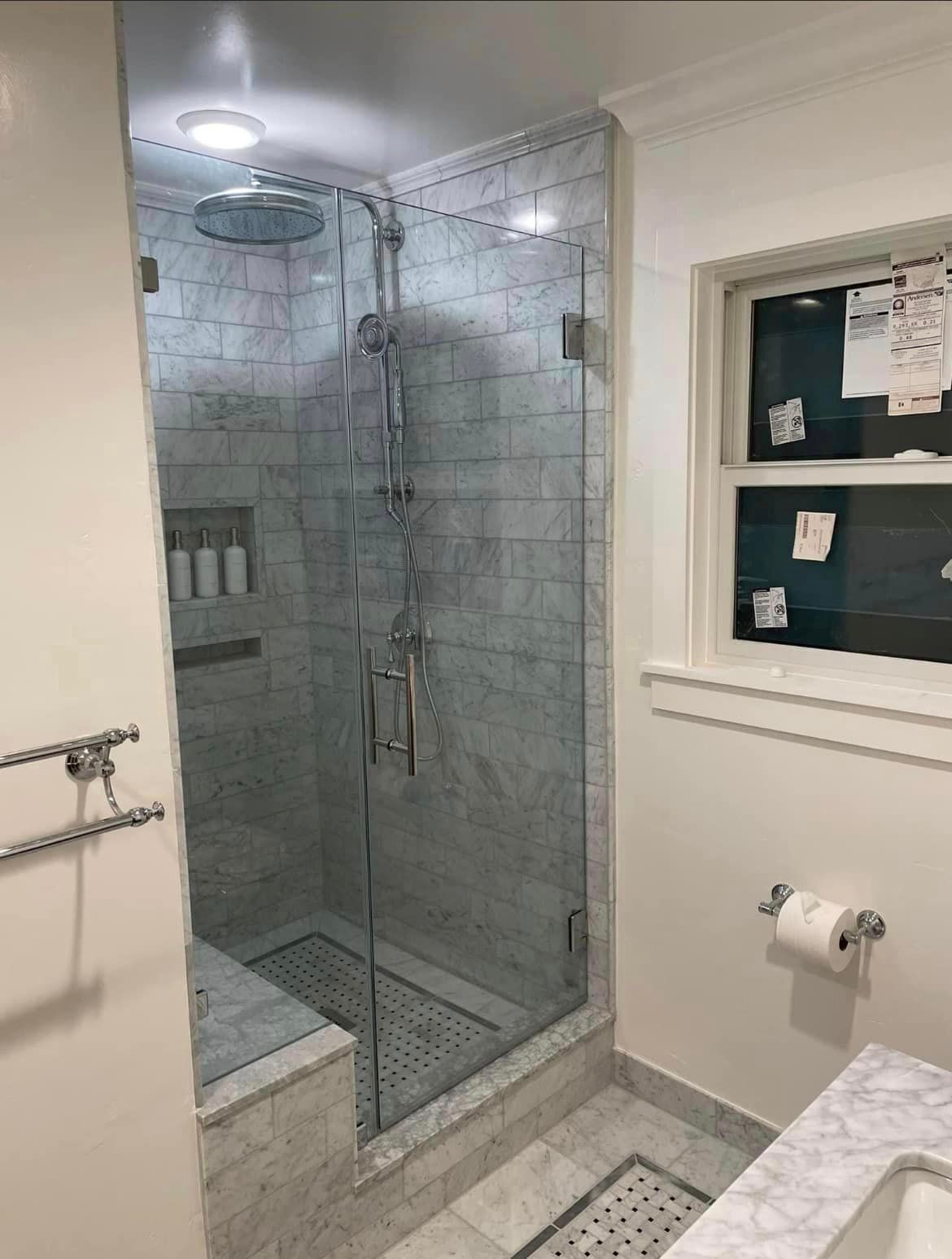 Walk-in shower with glass doors, tiled walls, and a built-in bench. A window is on the right.