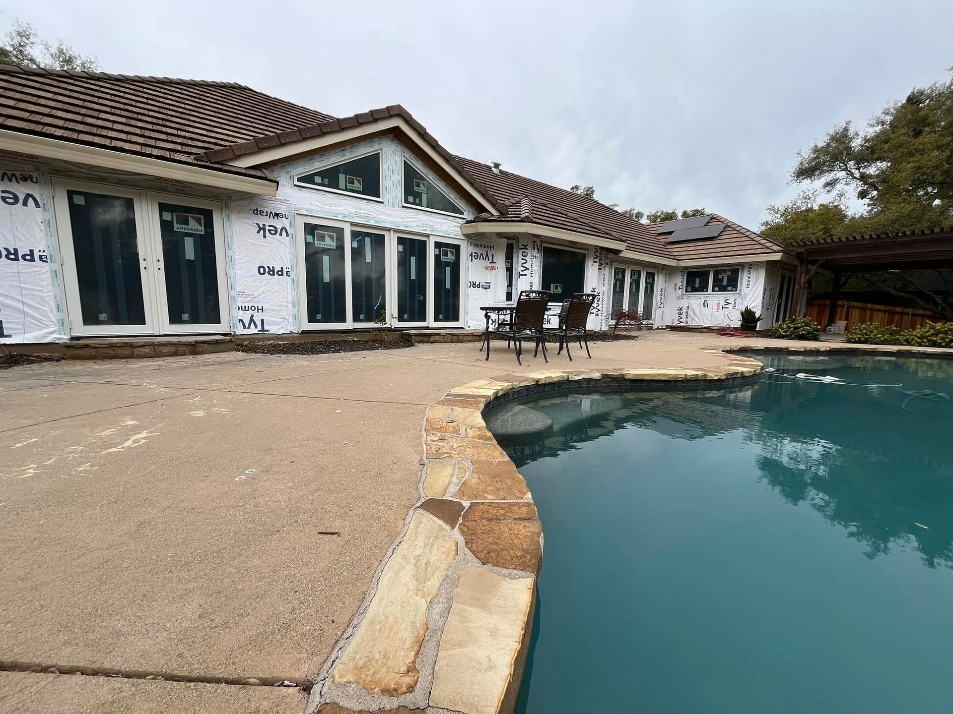 House exterior with pool under construction; white protective wrap on walls, brown tile roof, cloudy sky.