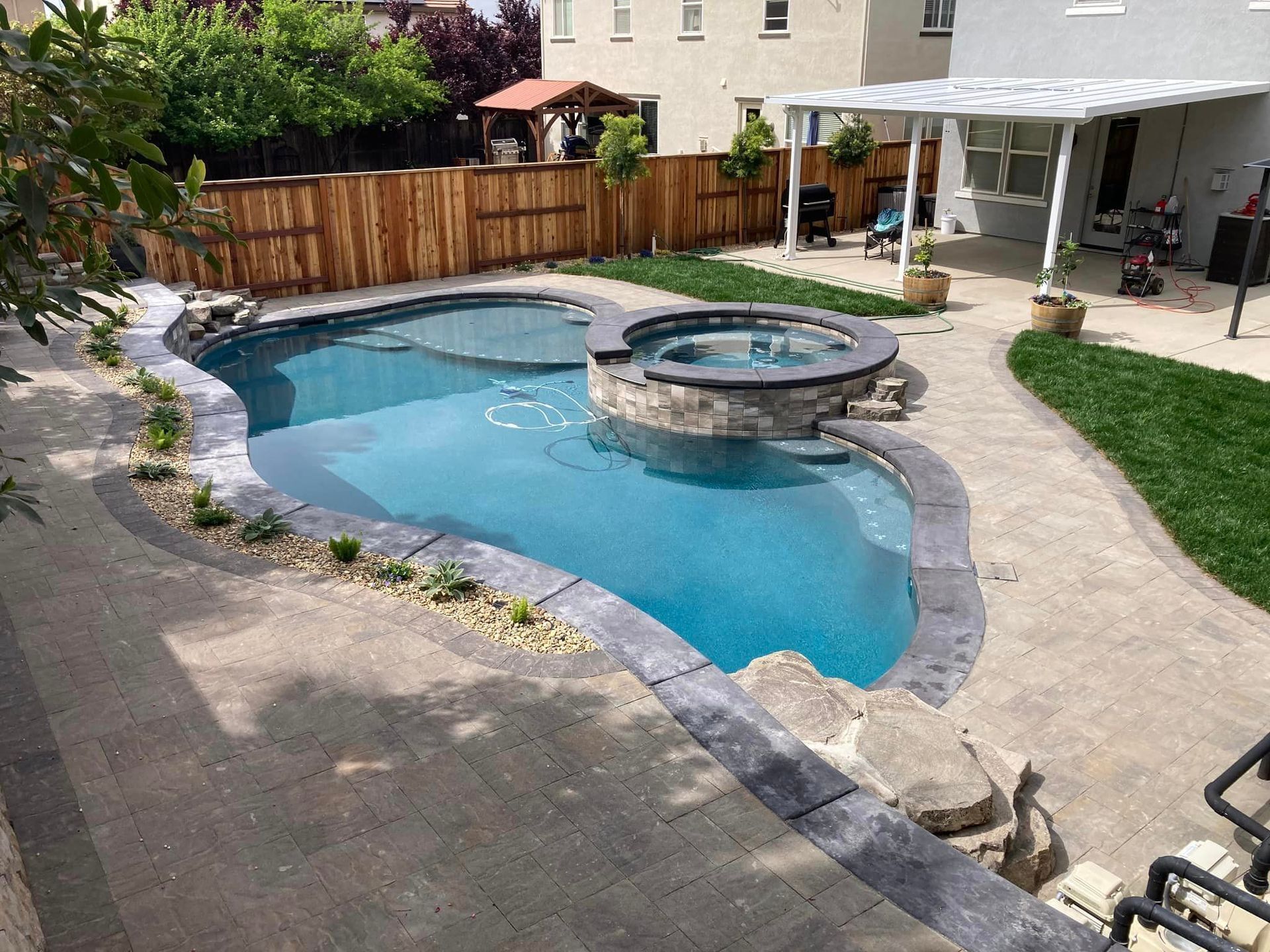Pool with a hot tub, surrounded by a patio and grass, wooden fence in the background.