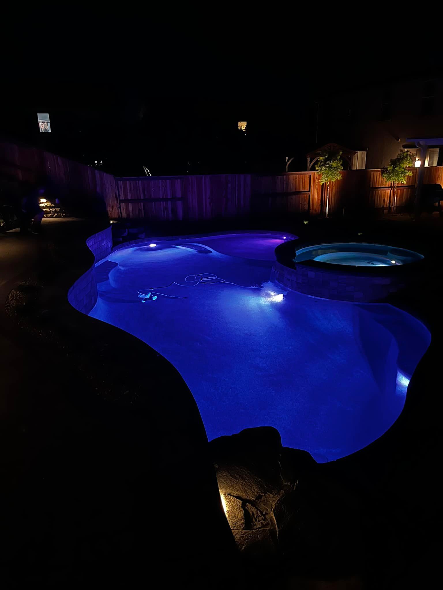 Pool at night with blue lights, next to a hot tub. Backyard setting.