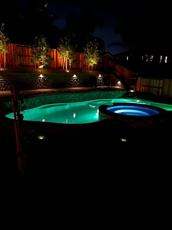 Nighttime view of a backyard pool and hot tub, illuminated with green and white lights. Trees and a wooden fence are also lit.