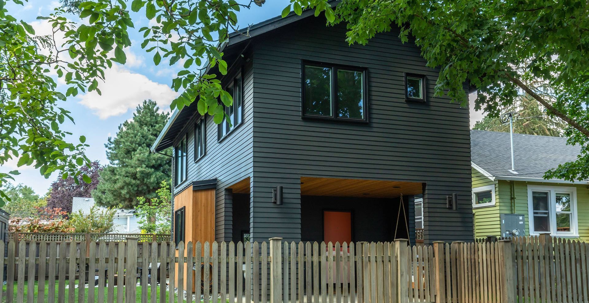 Modern two-story house with dark gray siding, orange door, and wooden fence in a residential area.