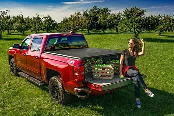 Red pickup truck with a woman sitting on the tailgate, crates of apples nearby, in an orchard.