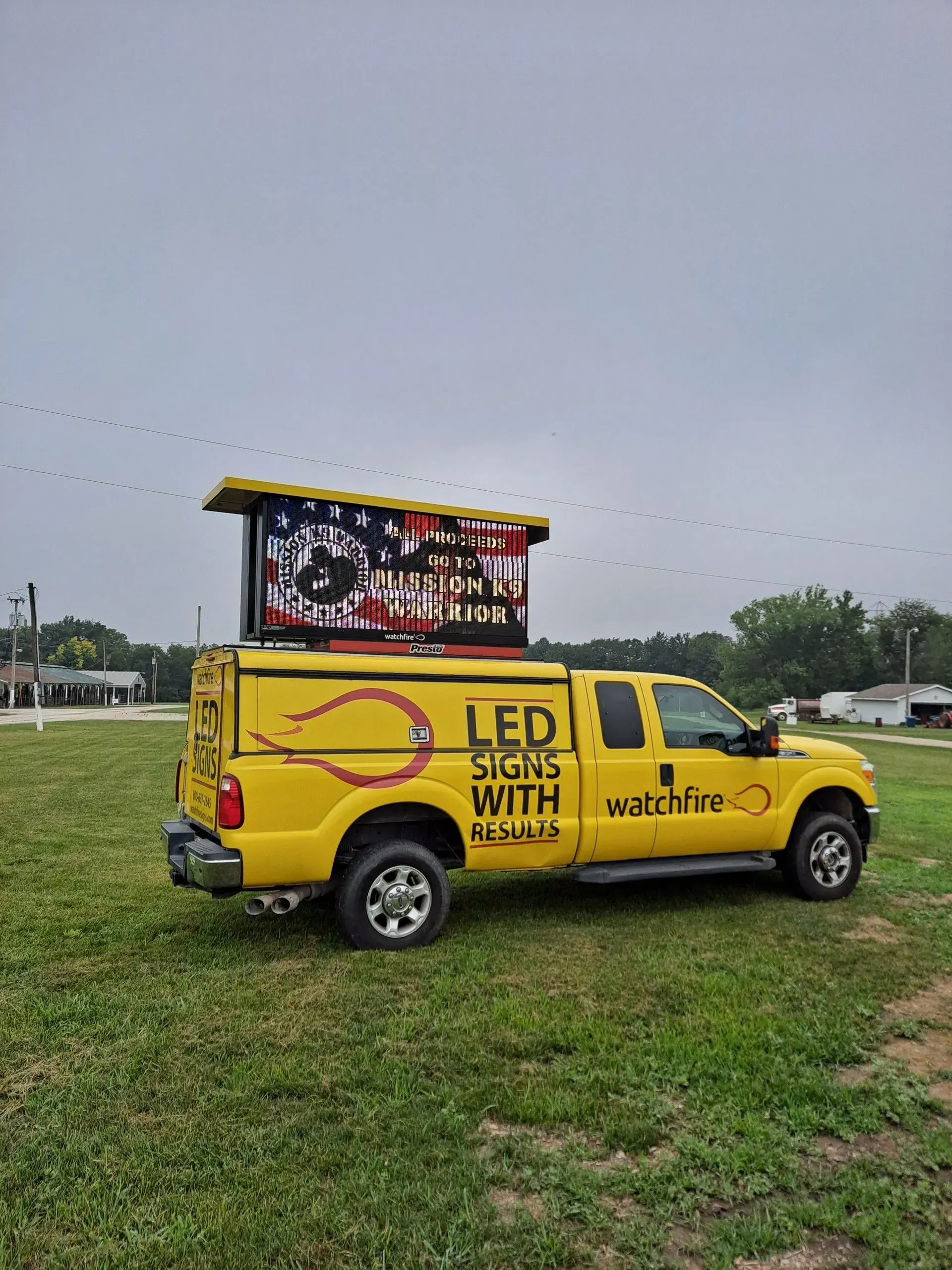 Yellow truck with LED sign display on a grassy field under overcast skies.