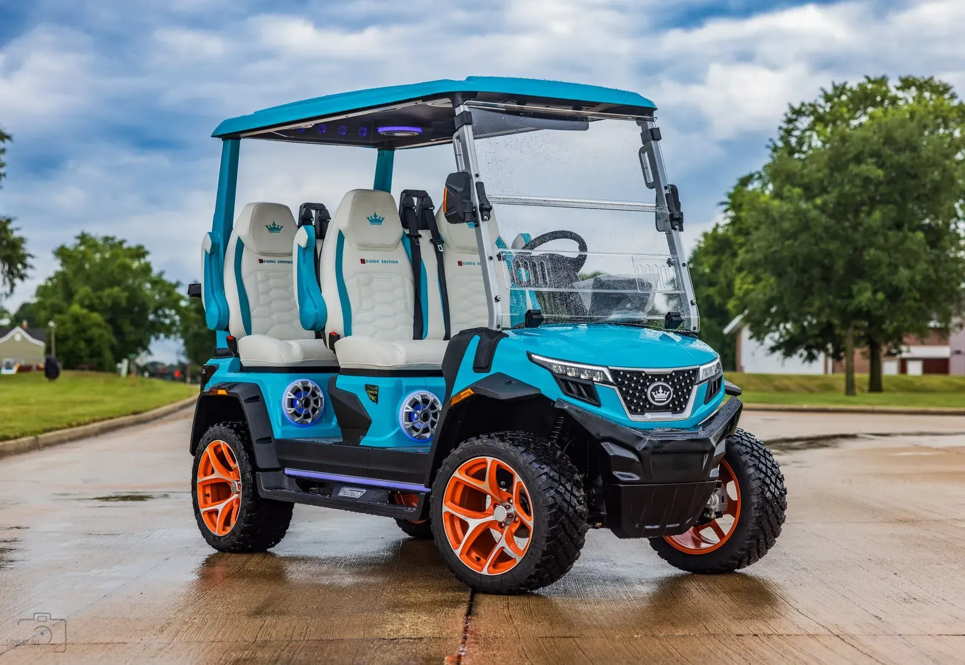 Teal and black custom golf cart with orange wheels on a paved road under a cloudy sky.