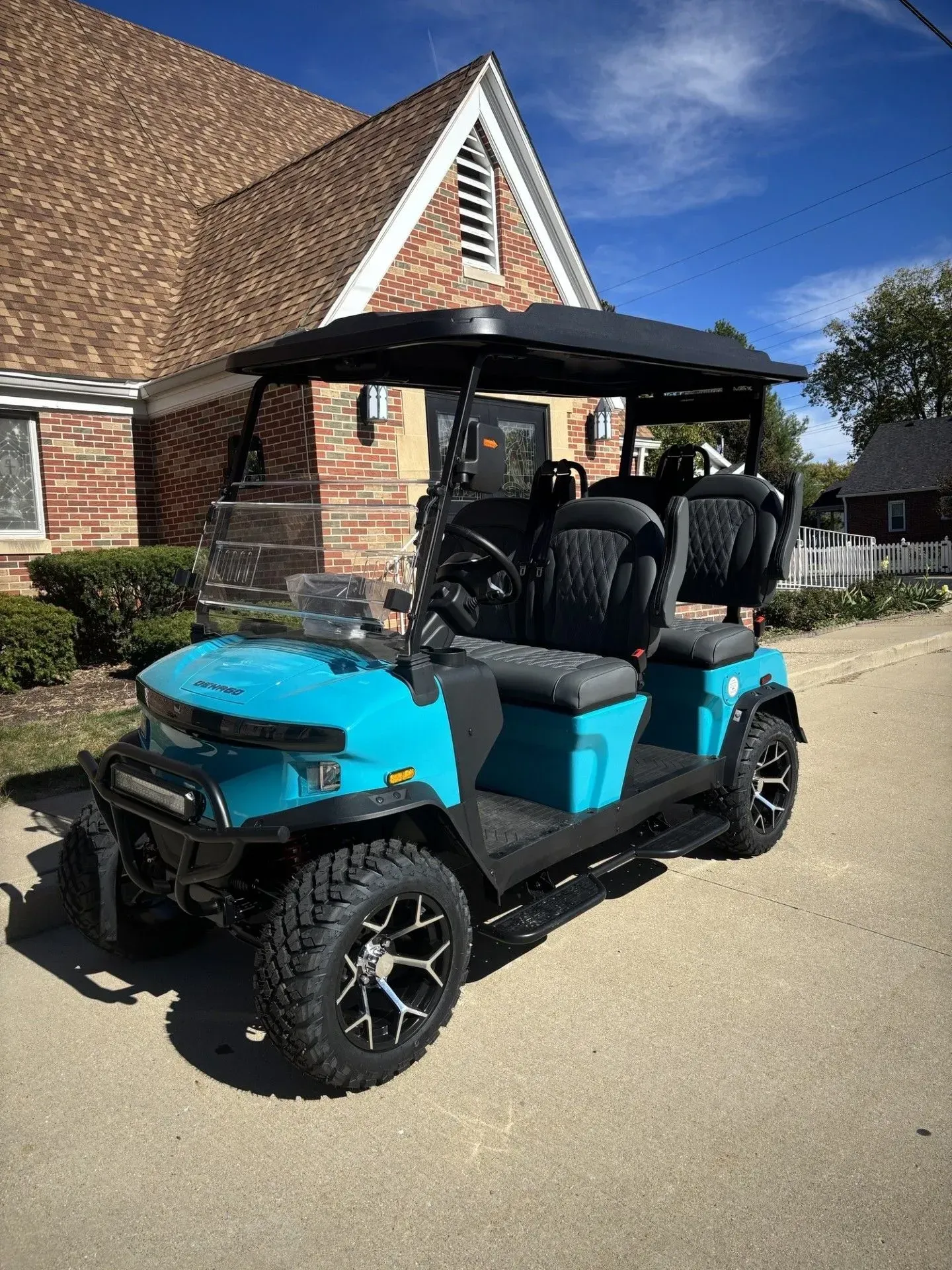 Turquoise golf cart with black roof and seats, parked in front of a brick building on a sunny day.