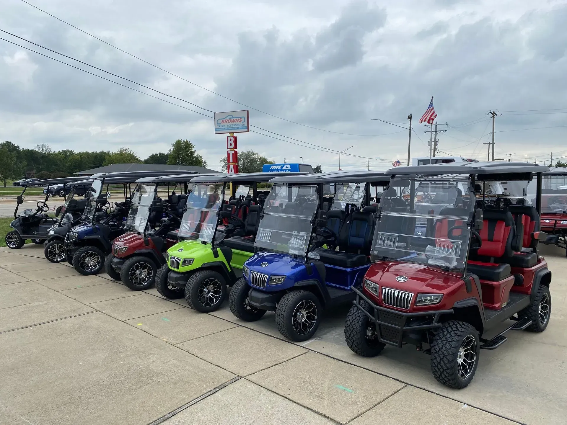 Row of colorful golf carts parked on pavement under a cloudy sky.