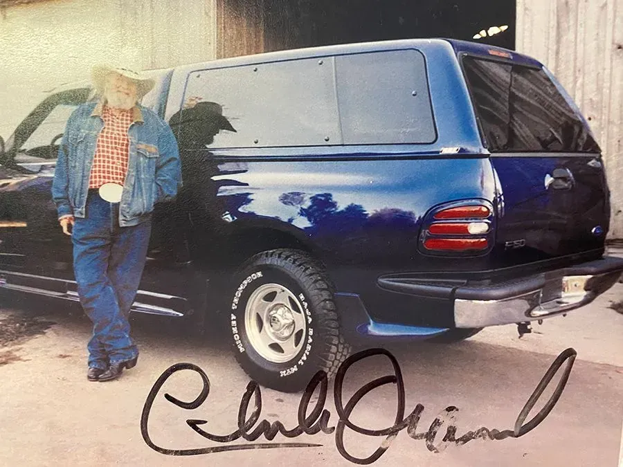 Man in cowboy attire stands next to a blue truck with a camper shell. Signature at the bottom.