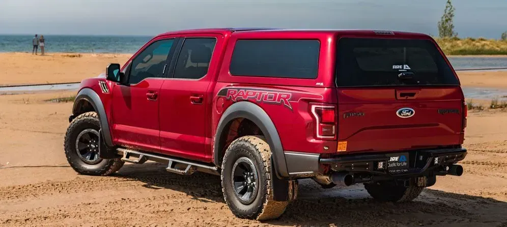 Red Ford Raptor pickup truck with a camper shell parked on a sandy beach.